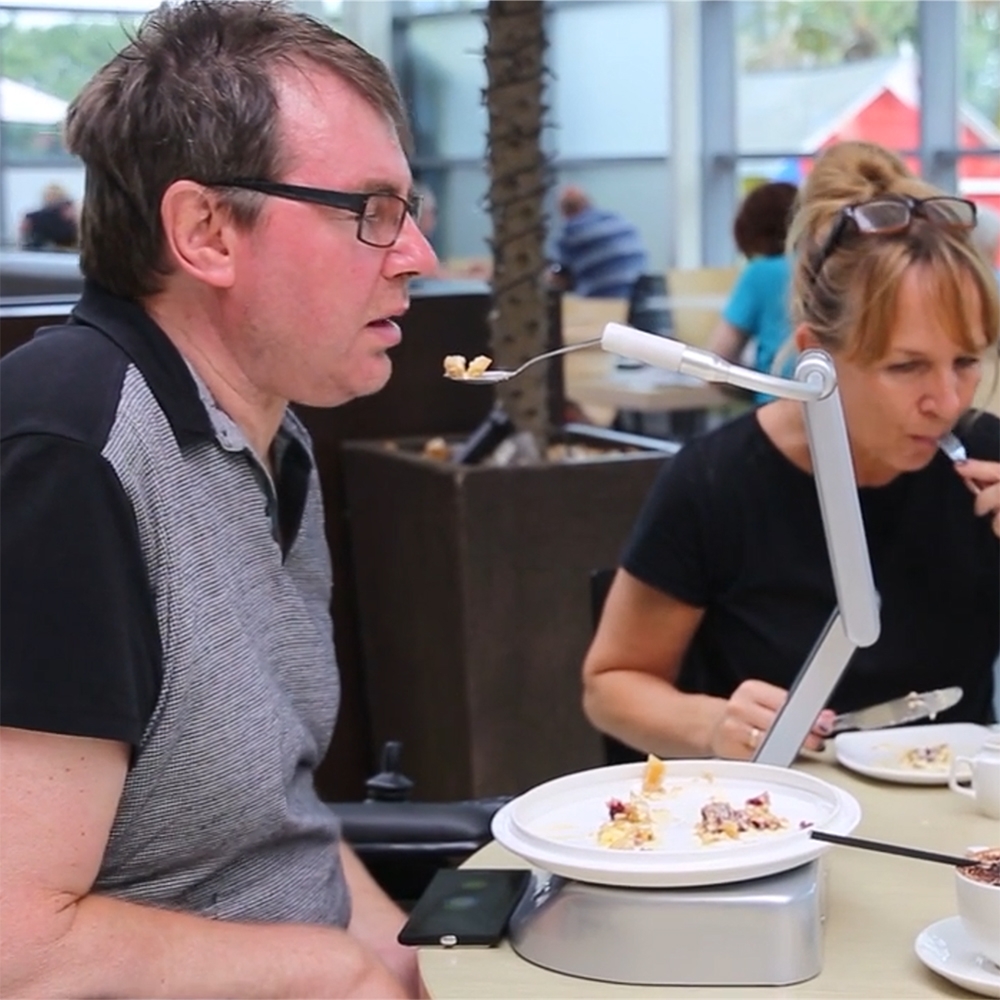 Man in glasses at a cafe table beside a robotic feeding arm holding a spoonful of food near his mouth